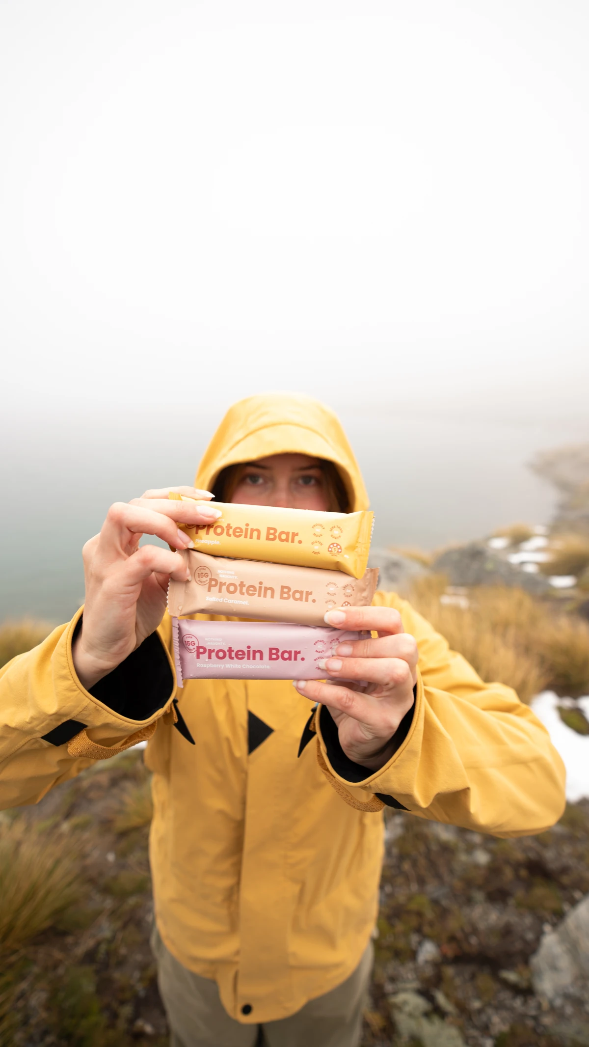 Three Nothing Naughty protein bars held towards the camera by a person in a yellow rain jacket